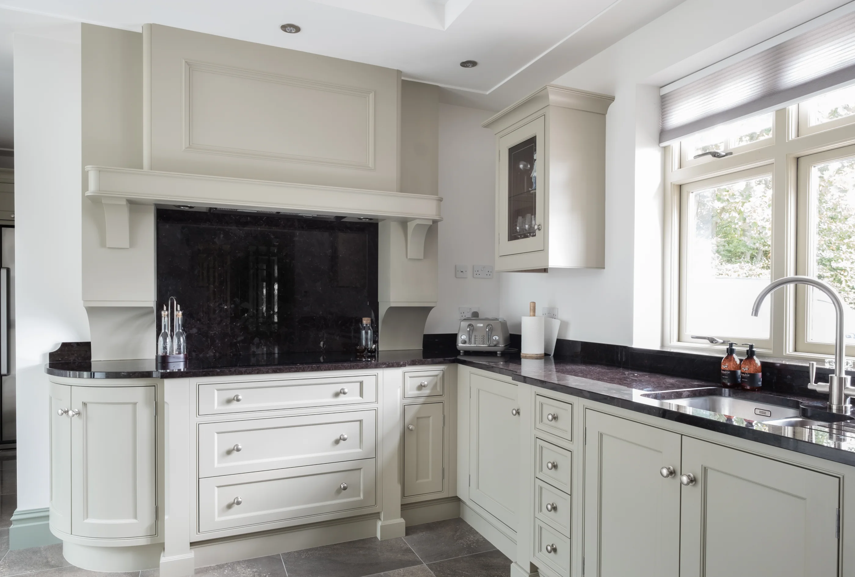 Pale fitted kitchen cabinetry arranged around a dark stone cooking centre and a window