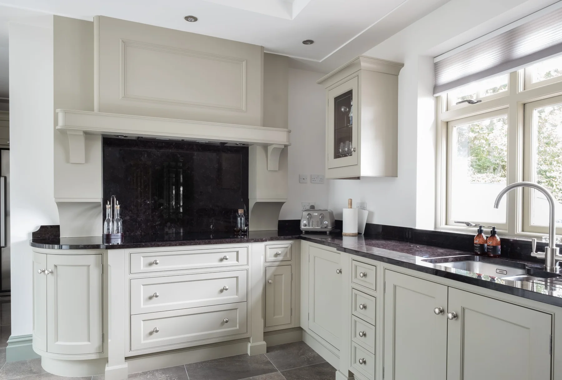 A pale fitted kitchen arranged around a dark stone fireplace