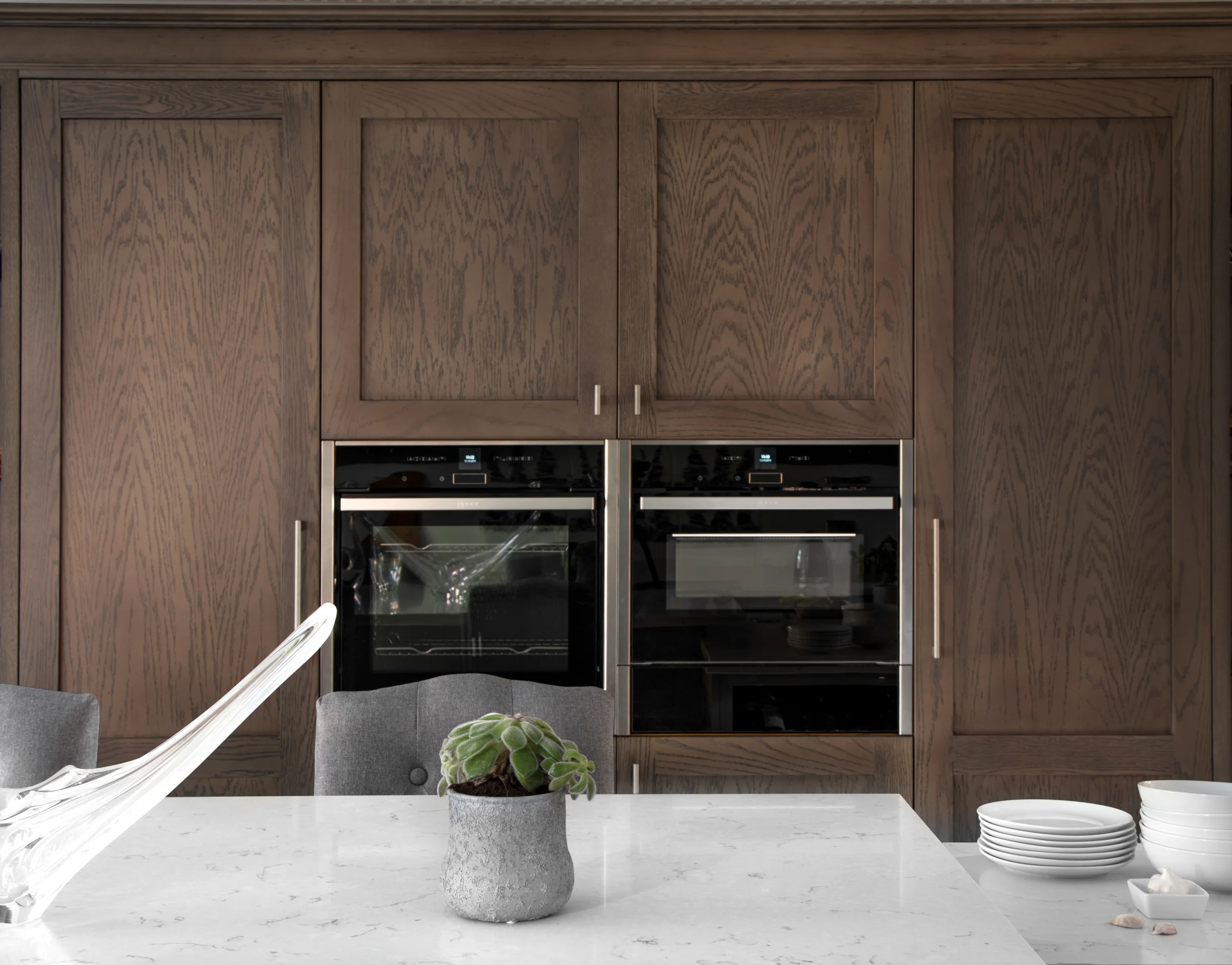 Dark oak kitchen cabinetry with integrated ovens behind a pale dining surface